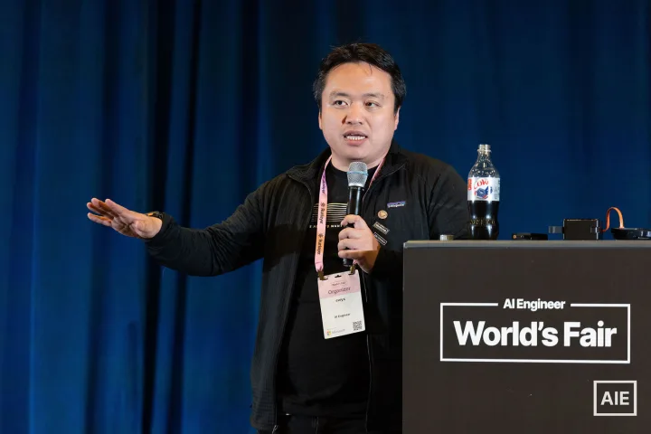 swyx speaking into a microphone against blue curtains at AI Engineer World’s Fair 2026
